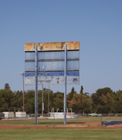 A large, weathered scoreboard stands tall in an open field with lush green grass in the foreground. The sky is clear and blue, while trees and a few structures are visible in the background. The rusty areas on the scoreboard indicate its age and exposure to the elements.