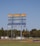 A large, weathered scoreboard stands tall in an open field with lush green grass in the foreground. The sky is clear and blue, while trees and a few structures are visible in the background. The rusty areas on the scoreboard indicate its age and exposure to the elements.