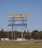 A large, weathered scoreboard stands tall in an open field with lush green grass in the foreground. The sky is clear and blue, while trees and a few structures are visible in the background. The rusty areas on the scoreboard indicate its age and exposure to the elements.