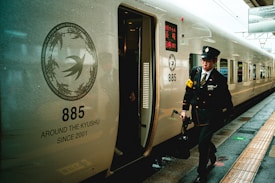A train conductor wearing a uniform and carrying a bag walks beside a sleek, modern train at a station. The train has the number 885 and the phrase 'Around the Kyushu since 2001' printed on its side. The platform is wet, suggesting recent rain.