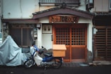 A traditional building with a wooden façade and sliding doors, featuring a wooden sign with Japanese characters above the entrance. A small motorcycle with a box attached is parked in front, and there is a covered bicycle or another vehicle to the left.