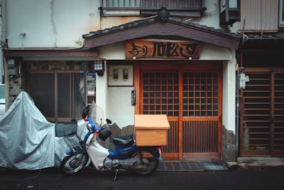 A traditional building with a wooden façade and sliding doors, featuring a wooden sign with Japanese characters above the entrance. A small motorcycle with a box attached is parked in front, and there is a covered bicycle or another vehicle to the left.