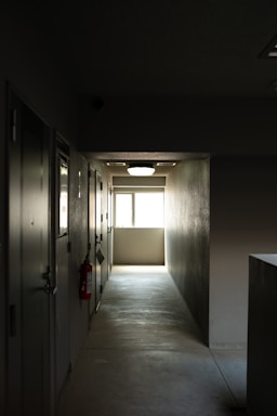 A dimly lit hallway with doors on the left, a bright window at the end, and a fire extinguisher mounted on the wall.