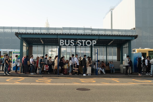 A crowded bus stop with a large group of people waiting in line. The shelter is modern with a glass and metal structure. Some individuals are seated on benches while others are standing and holding bags. Several buses are partially visible in the background.