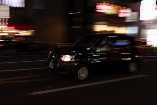 A sleek black taxi speeding through London streets at night with city lights glowing.