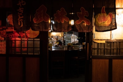 A dimly lit traditional Japanese restaurant with hanging banners featuring Japanese characters. The warm glow of interior lights illuminates a chef working behind a counter, surrounded by kitchen equipment and empty tables.