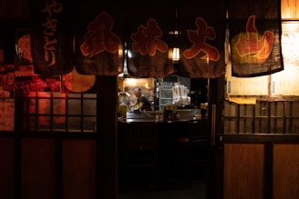 A dimly lit traditional Japanese restaurant with hanging banners featuring Japanese characters. The warm glow of interior lights illuminates a chef working behind a counter, surrounded by kitchen equipment and empty tables.