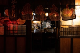 A dimly lit traditional Japanese restaurant with hanging banners featuring Japanese characters. The warm glow of interior lights illuminates a chef working behind a counter, surrounded by kitchen equipment and empty tables.