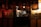A dimly lit traditional Japanese restaurant with hanging banners featuring Japanese characters. The warm glow of interior lights illuminates a chef working behind a counter, surrounded by kitchen equipment and empty tables.