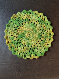 A circular crocheted doily with a scalloped edge. The design features alternating bands of green and yellow yarn, creating a gradient effect. The doily is intricately patterned with small gaps throughout, showcasing a delicate and detailed craftsmanship. It is placed on a textured dark surface.