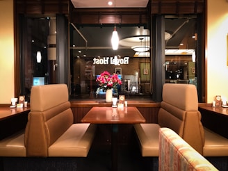 A cozy restaurant interior with beige upholstered booth seating around a wooden table. A window with decorative lettering offers a view into another part of the restaurant. A vase with vibrant red and pink flowers serves as a centerpiece on the table. Ambient lighting from modern pendant lamps creates a warm and inviting atmosphere.