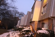 A row of townhouses with snow-covered shrubs in front. The sky is clear with a bluish tint, and trees without leaves are visible in the background. Warm light emanates from some windows, casting a cozy glow on the exterior.