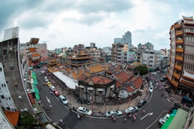 A bustling urban scene depicting a traditional Asian temple with elaborate, ornate roofs surrounded by a dense cluster of modern buildings. The street is busy with pedestrians, and several cars are parked along the roads that encircle the temple. The architecture of the temple contrasts with the surrounding cityscape, contributing to an eclectic and vibrant atmosphere.