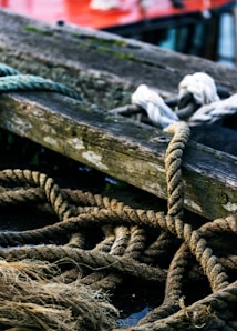 A weathered wooden dock with several thick, coiled ropes lying on it. The wood shows signs of moss and age, and the ropes are frayed, indicating heavy use. In the background, there is a hint of a red structure, possibly part of a boat.