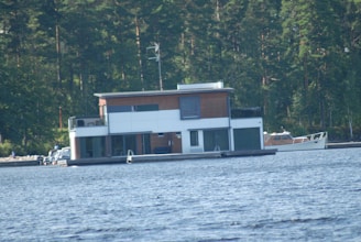 A modern two-story houseboat is floating on a lake, surrounded by dense forest. The houseboat features large windows and a flat roof. Nearby, a small boat is moored on the lake.