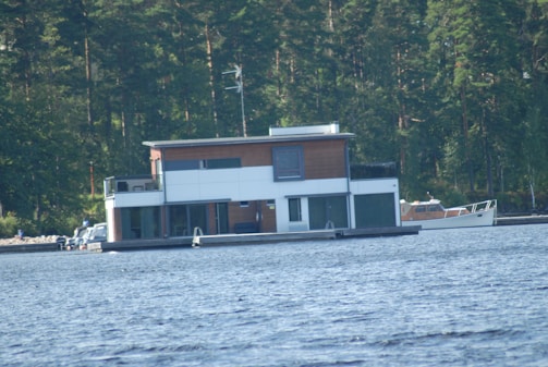 A modern two-story houseboat is floating on a lake, surrounded by dense forest. The houseboat features large windows and a flat roof. Nearby, a small boat is moored on the lake.
