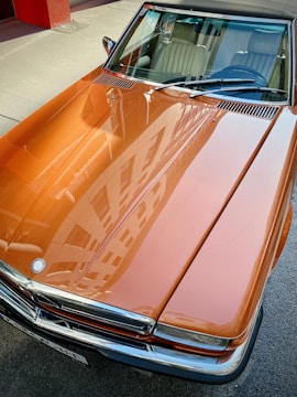 A polished, orange vintage car with a reflective surface showing the sky and surrounding buildings. The car's hood and windshield are prominently visible, displaying sleek lines and chrome details.