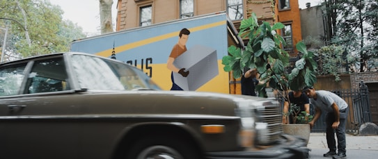 A vintage car is parked on a street with an urban backdrop, including brownstone buildings. In the background, a moving truck with a large graphic of a person carrying a box is visible. People are tending to a large potted plant, possibly relocating it from the truck or arranging it in its spot.