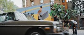 A vintage car is parked on a street with an urban backdrop, including brownstone buildings. In the background, a moving truck with a large graphic of a person carrying a box is visible. People are tending to a large potted plant, possibly relocating it from the truck or arranging it in its spot.
