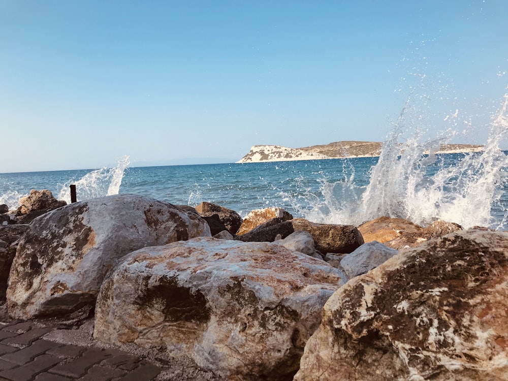 brown rocky shore with ocean waves crashing on rocks under blue sky during daytime