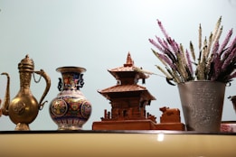 A collection of decorative items is displayed on a shelf. From left to right, there is an ornate golden teapot, a colorful ceramic vase with intricate patterns, a wooden miniature of a traditional Asian temple, and a metal bucket filled with artificial lavender flowers. The background is a pale blue, creating a serene atmosphere.