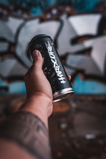 A person's tattooed arm is holding a black spray paint can with the word 'Hardcore' printed on it. The background shows a blurred graffiti-covered wall, giving a sense of urban art.