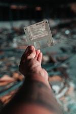 A hand is holding a vintage floppy disk against a blurred background with a bokeh effect. The floppy disk appears worn and aged, suggesting it has been used frequently or stored for a long time.