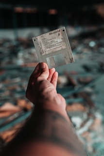 A hand is holding a vintage floppy disk against a blurred background with a bokeh effect. The floppy disk appears worn and aged, suggesting it has been used frequently or stored for a long time.
