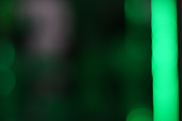 A blurred abstract image primarily featuring shades of green with a prominent bright vertical stripe on the right side. The background consists of dark green and soft bokeh circles creating a soft and diffuse appearance.