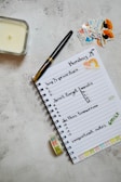 A neatly arranged journal or planner lays open on a textured surface. It has handwritten notes listing 'top 3 priorities', 'don't forget meals', 'do this tomorrow', and 'important notes'. A black and gold pen rests on the open page. Nearby are decorative stickers with colorful designs. A candle is placed to the left and a roll of washi tape lies near the notebook.