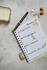 A neatly arranged journal or planner lays open on a textured surface. It has handwritten notes listing 'top 3 priorities', 'don't forget meals', 'do this tomorrow', and 'important notes'. A black and gold pen rests on the open page. Nearby are decorative stickers with colorful designs. A candle is placed to the left and a roll of washi tape lies near the notebook.