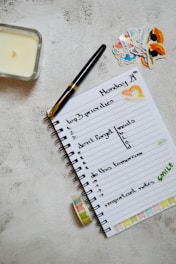 A neatly arranged journal or planner lays open on a textured surface. It has handwritten notes listing 'top 3 priorities', 'don't forget meals', 'do this tomorrow', and 'important notes'. A black and gold pen rests on the open page. Nearby are decorative stickers with colorful designs. A candle is placed to the left and a roll of washi tape lies near the notebook.