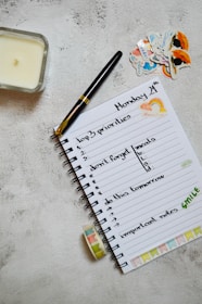A neatly arranged journal or planner lays open on a textured surface. It has handwritten notes listing 'top 3 priorities', 'don't forget meals', 'do this tomorrow', and 'important notes'. A black and gold pen rests on the open page. Nearby are decorative stickers with colorful designs. A candle is placed to the left and a roll of washi tape lies near the notebook.