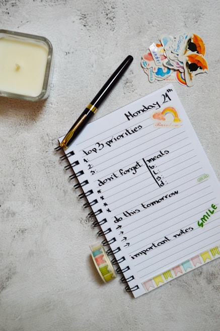 A neatly arranged journal or planner lays open on a textured surface. It has handwritten notes listing 'top 3 priorities', 'don't forget meals', 'do this tomorrow', and 'important notes'. A black and gold pen rests on the open page. Nearby are decorative stickers with colorful designs. A candle is placed to the left and a roll of washi tape lies near the notebook.