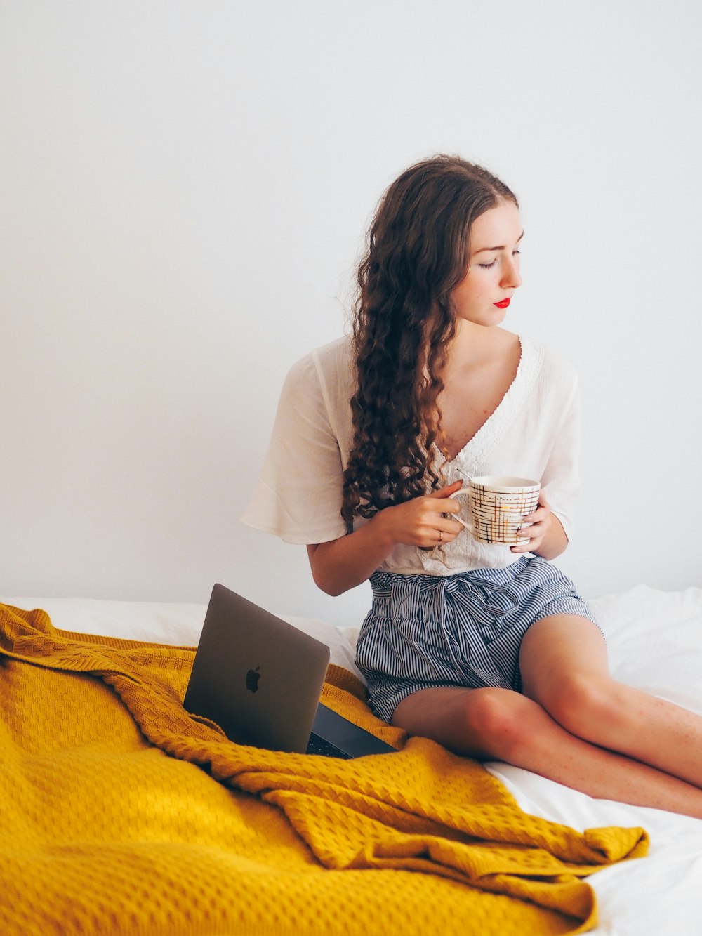 Frau in weißem Langarmhemd und blauen Jeansshorts sitzt mit silbernem MacBook auf dem Bett