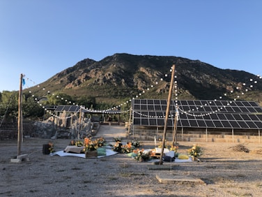 A picturesque outdoor setting with a mountainous backdrop. Solar panels are strategically placed in the foreground, and string lights add a decorative element. Festive floral arrangements with vibrant orange flowers and cushions are set on a mat, suggesting a prepared gathering area.