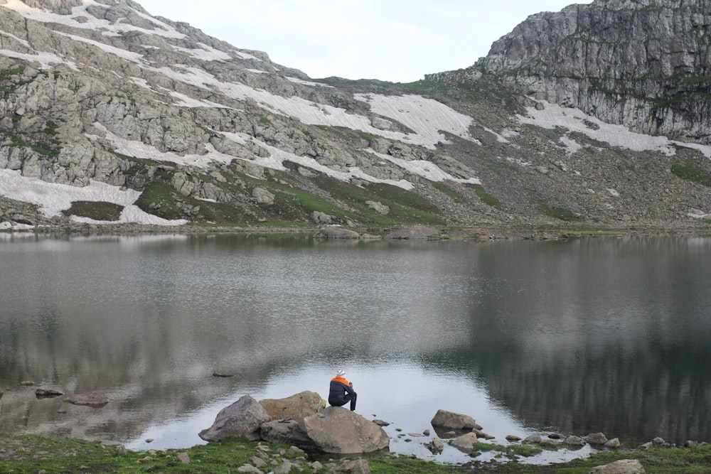 person in orange jacket sitting on rock near lake during daytime