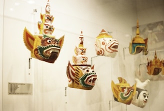 Several ornate masks are displayed in a glass case. These masks feature elaborate designs with bold facial expressions, prominent eyes, and wide, open mouths. The masks appear to have traditional cultural significance, possibly representing mythological characters. They vary in colors and intricate patterns.