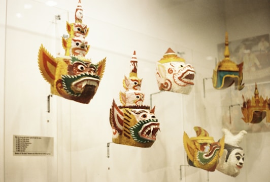 Several ornate masks are displayed in a glass case. These masks feature elaborate designs with bold facial expressions, prominent eyes, and wide, open mouths. The masks appear to have traditional cultural significance, possibly representing mythological characters. They vary in colors and intricate patterns.