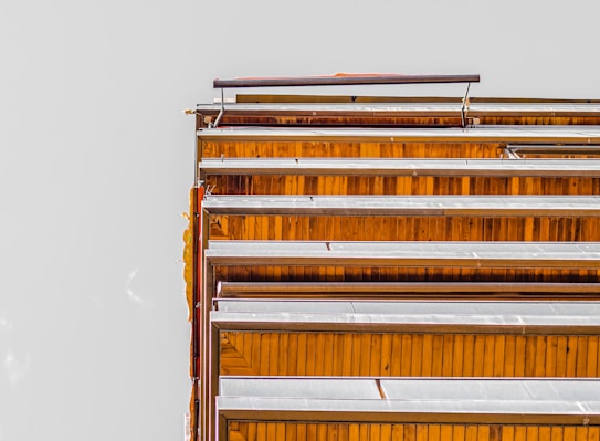 A modern architectural structure featuring an arrangement of wooden and metallic horizontal slats, set against a plain background. The slats have a repetitive, geometric pattern.