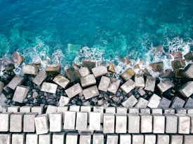 Aerial view of a coastal area where the ocean meets a structured shoreline composed of large, geometric concrete blocks. The blue and teal ocean waves gently crash against the blocks, creating white foam.