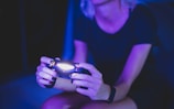 A person with short hair is holding a lighted game controller in a dimly lit room, creating an atmosphere with a predominant purple and blue hue. Their focus appears to be on the gaming activity.