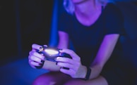 A person with short hair is holding a lighted game controller in a dimly lit room, creating an atmosphere with a predominant purple and blue hue. Their focus appears to be on the gaming activity.