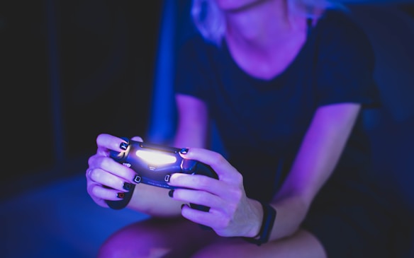 A person with short hair is holding a lighted game controller in a dimly lit room, creating an atmosphere with a predominant purple and blue hue. Their focus appears to be on the gaming activity.