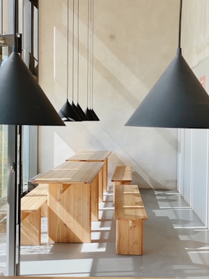 A modern interior space featuring a set of wooden tables and benches positioned in a sunlit room. Black pendant lamps with cone-shaped shades are suspended from the ceiling, casting geometric shadows on the walls and floor. The minimalist design and natural lighting create a serene atmosphere.