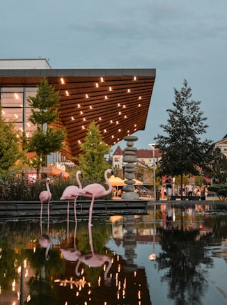 A modern architectural structure with a wooden overhang brightly lit by numerous small lights reflects off a nearby water body. Several pink flamingo statues are placed in the shallow water, along with green trees and a stack of round stones. People and market stalls can be seen in the background, creating a lively atmosphere.