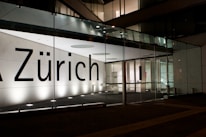 A modern building facade featuring large glass panels and a brightly illuminated interior. The word 'Zürich' is displayed prominently across the glass. The interior appears minimalist and well-lit, with visible structural elements and reflections on the glass surface.