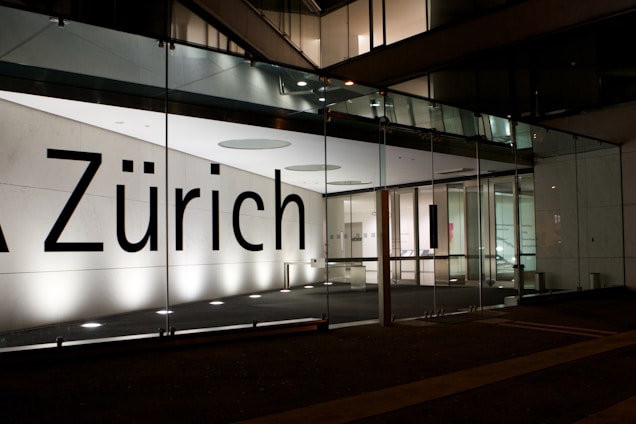 A modern building facade featuring large glass panels and a brightly illuminated interior. The word 'Zürich' is displayed prominently across the glass. The interior appears minimalist and well-lit, with visible structural elements and reflections on the glass surface.