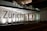 A modern building facade featuring large glass panels and a brightly illuminated interior. The word 'Zürich' is displayed prominently across the glass. The interior appears minimalist and well-lit, with visible structural elements and reflections on the glass surface.