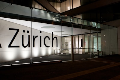 A modern building facade featuring large glass panels and a brightly illuminated interior. The word 'Zürich' is displayed prominently across the glass. The interior appears minimalist and well-lit, with visible structural elements and reflections on the glass surface.